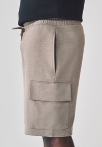Pantaloncini sportivi beige con vita elastica, tasca laterale e una grande tasca applicata sulla gamba destra; tessuto liscio e leggero.