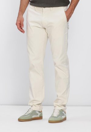 Man draagt beige broek en lichtgroene sneakers, staand met handen in de zakken tegen een effen achtergrond.