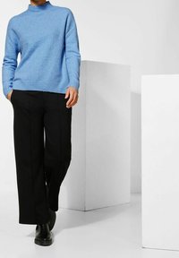 Helleblaue gestrickte Sweater mit einem hohen Kragen, kombiniert mit schwarzen weit geschnittenen Hosen und schwarzen Stiefeln, vor einem minimalistischen Hintergrund.