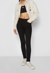 Mujer vistiendo una chaqueta beige, top negro de corte corto, jeans skinny negros de cintura alta y zapatillas blancas, de pie contra un fondo blanco.