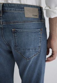 Blue denim jeans featuring a leather logo patch at the waistband, contrasted stitching, and a back pocket with a subtle curved design.