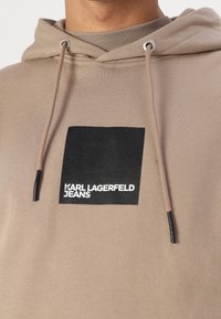 Brun hættetrøje med snor i hætten og et sort firkantet mærke med "KARL LAGERFELD JEANS" i hvid tekst. Blød tekstur, afslappet design.
