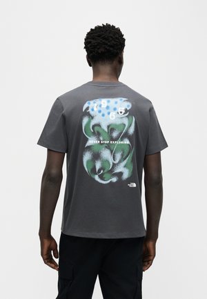 Άνδρας που φοράει σκούρο γκρι μπλουζάκι με πράσινο, μπλε και λευκό αφηρημένο σχέδιο, αριθμούς και το κείμενο "ΜΗ ΣΤΑΜΑΤΑΣ ΠΟΤΕ ΝΑ ΕΞΕΡΕΥΝΑΣ" στην πλάτη.