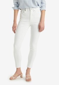 Bílé skinny džíny s vysokým pasem vyrobené z denimu, s úzkým střihem a předními kapsami, stylizované s béžovými sandály na blokovém podpatku.