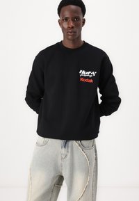 Sort sweatshirt med "Huf Racing" og "Kodak" tryk i hvid og orange, med rund hals og lange ærmer. Båret med lyseblå slidte jeans.