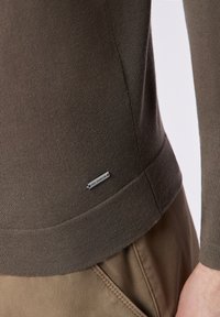 Brauner, taillierter Pullover mit glatter Textur. Verfügt über einen gerippten Saum und ein kleines silbernes Etikett mit dem Markennamen an der unteren Seite.