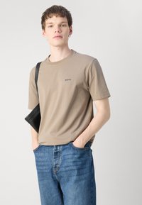 Beige bomull t-skjorte med korte ermer og rund hals, med en liten svart "BOSS"-logo på brystet; parret med blå jeans.