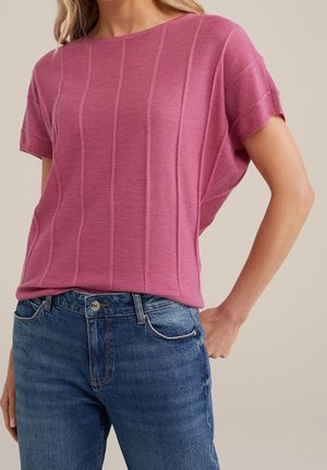 Vrouw die een roze gebreide top met korte mouwen en verticale ribbels draagt, ingestopt in een hoog uitgesneden blauw spijkerbroek, tegen een neutrale achtergrond.