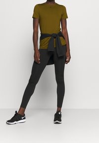 Camisa de manga corta verde oliva con leggings negros, tela negra atada en la cintura, combinada con zapatillas deportivas negras, textura suave.