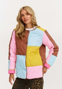 Žena s dlhými blond vlasmi má na sebe prešívanú patchworkovú bundu v hnedých, modrých, žltých, ružových farbách a nohavice s leopardím vzorom, stojí na bielom pozadí.