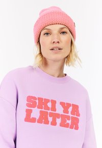 Sudadera de color púrpura claro con "SKI YA LATER" en letras rojas texturizadas; combinada con un gorro de punto rosa con puño acanalado.