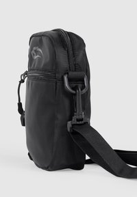 Zwarte nylon schoudertas met een hoofdvak met rits, verstelbare riem en een ronde logodetail aan de voorkant. Soepele textuur.