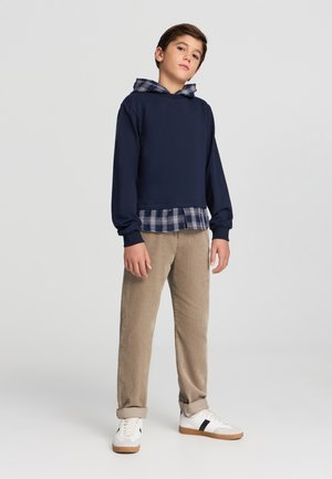 Felpa navy con accenti blu a quadri, abbinata a pantaloni in corduroy beige e sneakers bianche con strisce nere. Look casual a strati.