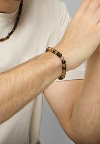 Pulsera de cuentas hecha de piedras marrones, negras y tostadas con acentos de patrón. Lleva puesta en una muñeca con cabello claro y piel del antebrazo visible.