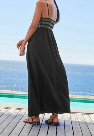 Vestido largo negro con un cuerpo fruncido, tirantes finos y una silueta fluida. Lleva sandalias negras con tiras; el fondo presenta un mar azul.