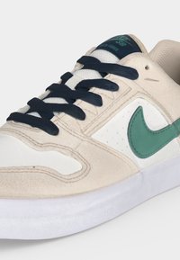 Μπεζ αθλητικό παπούτσι Nike SB Delta Force με темноμπλε κορδόνια, τιρκουάζ λογότυπο Nike swoosh και λευκή σόλα, γωνιακά για να δείχνει λεπτομέρειες εμπρός και πλάι.