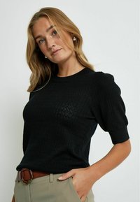 Pull en tricot noir avec des manches courtes bouffantes, des motifs texturés, des poignets côtelés et un col rond. Assorti à un pantalon beige.