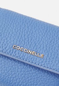 Modra usnjena torbica z zrnato teksturo, ki ima zlat logotip "CoCCinelle" in kontrastne šive vzdolž robov.