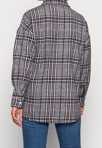 Persoon in een zwart, wit en rood geruit overhemd met lange mouwen, gecombineerd met blauwe jeans, gezien vanaf de achterkant.