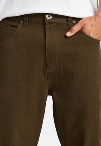 Bruine katoenen broek met een gestructureerde afwerking, voorzien van een voorzak, knoopsluiting en riemlussen, gepresenteerd tegen een witte achtergrond.