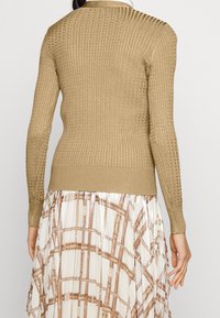Pletený svetřík ve světle hnědé barvě s žebrovanou texturou, dlouhými rukávy, kulatým výstřihem a úzkými manžetami, kombinovaný s vzorovanou sukní.