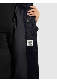 Mornarsko plava termo jakna s glatkom, sjajnom vanjskom površinom. Ima unutarnji džep na zatvarač i oznaku na kojoj piše "Topli zagrljaj od tkanine."