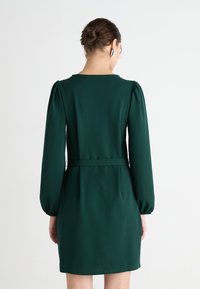 Zelené šaty s vysokým golierom, naberanými dlhými rukávmi a opaskom v páse. Vyrobené z hladkého materiálu s dĺžkou po strednú časť stehna.