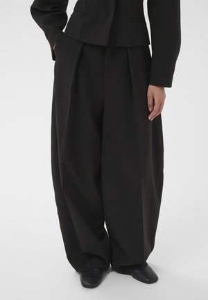 Personne vêtue d'un pantalon plissé noir ample et d'une veste noire assortie, debout avec une main dans la poche et des chaussures noires visibles.