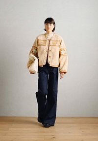 Beżowa polarowa kurtka z wielokolorowymi paskami, drewnianymi guzikami i szerokimi rękawami, w połączeniu z ciemnoniebieskimi spodniami z wysokim stanem i beżową torbą.