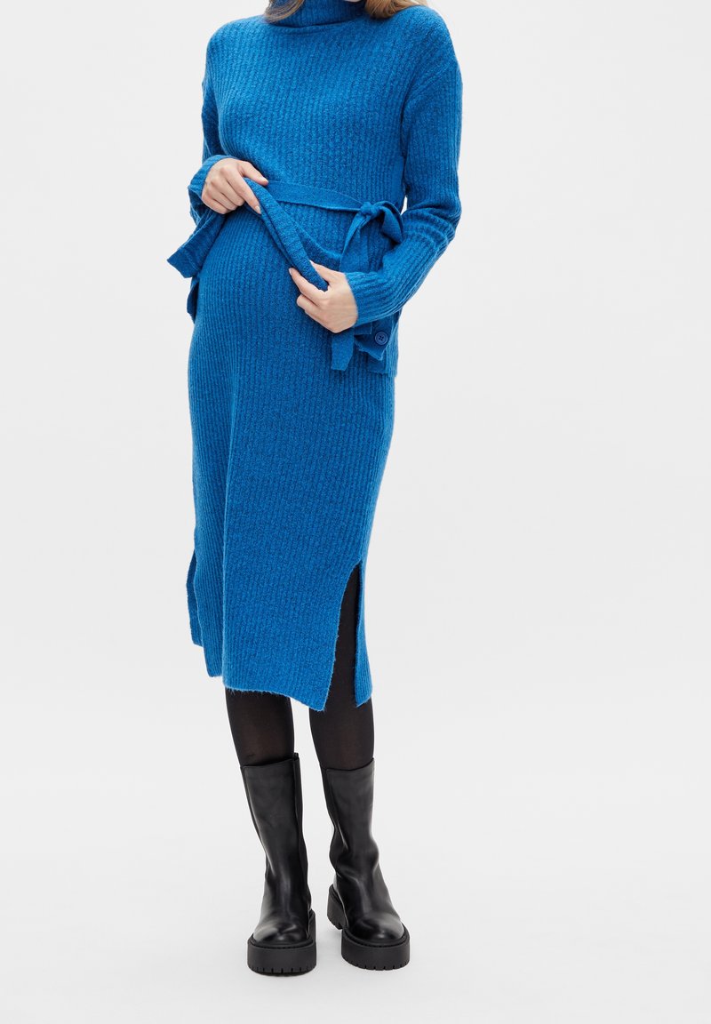 Robe en maille bleu vif, avec un col roulé, une taille ceinturée, des fentes latérales et une texture côtelée. Portée avec des bottes noires montantes jusqu'au genou.