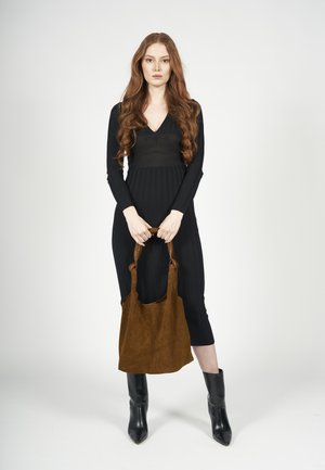 Schwarzes, geripptes Strickkleid mit tiefem V-Ausschnitt, kombiniert mit einer braunen Wildleder-Tote. Das Model trägt schwarze spitze Stiefeletten und steht zentral.