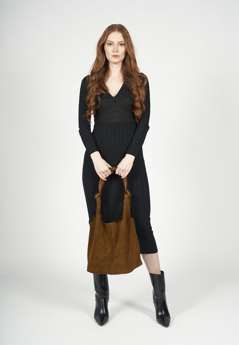 Zwart geribbeld gebreid jurkje met een diepe V-hals, gecombineerd met een bruine suède tote bag. Model draagt zwarte puntige enkel laarzen en staat centraal.