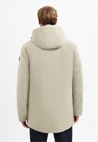 Beige gewatteerde jas met een capuchon, voorzien van een zachte textuur en een iets langere achterkant voor extra bedekking. Minimalistisch ontwerp zonder zichtbare details.