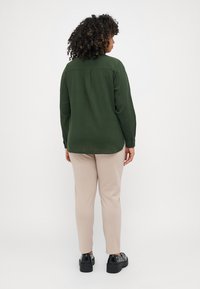 Zelená košile na knoflíky s texturovanou tkaninou, dlouhými rukávy a záhybem na zádech, spárovaná s béžovými slim-fit kalhotami a černými platformovými botami.