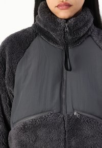 Giacca in pile grigia con zip, caratterizzata da un colletto alto, un tessuto morbido e a trama, e due tasche frontali con zip dotate di pannelli a contrasto.