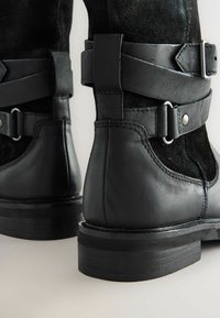 Stivali alla caviglia in pelle e suede neri con dettagli in fibbia e cinturino, caratterizzati da una suola piatta e punta rotonda, che mettono in evidenza texture in contrasto.