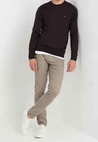 Maglione a maglia marrone scuro con scollatura rotonda e piccolo logo, abbinato a pantaloni chinos beige e sneakers bianche. Design semplice e aderente.
