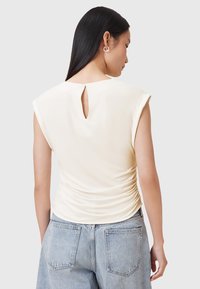 Crèmekleurige mouwloze top met een hoge halslijn, een sleutelgatdetail aan de achterkant en ruches aan de zijkanten, gecombineerd met lichtblauwe denim jeans.