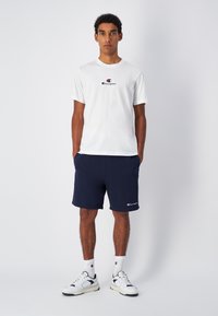Weißes Baumwoll-T-Shirt mit schwarzem und rotem Champion-Logo, kombiniert mit navy Shorts, die ein Logo auf dem linken Bein haben und weißen Sneakers.