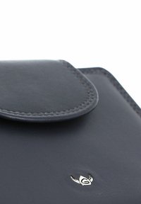 Coin de portefeuille en cuir noir avec rabat cousu et petit logo métallique argenté en forme d'emblème ailé stylisé.