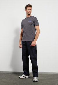 Grå t-skjorte med rund hals og lilla grafisk trykk, kombinert med mørkeblå denimjeans og sølvfargede sportssko, stående på et grått gulv.