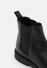 Botte Chelsea en cuir noir avec panneaux latéraux élastiques, languette de tirage et semelle en caoutchouc texturée. Finition lisse avec un minimum de détails de couture.