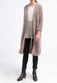 Lång, öppen kofta i mjuk beige mohairblandning, lagd över en strukturerad vit topp och mönstrad blus, ihop med svarta skinny jeans och ankelskoboots.