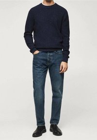 Mężczyzna w ciemnogranatowym swetrze z melanżem, niebieskich dżinsach i czarnych skórzanych butach, stoi z jedną ręką w kieszeni na jednolitym tle.