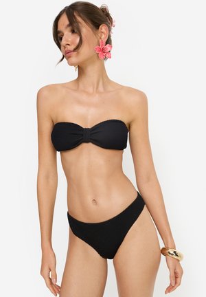Woman wearing black textured bandeau bikini with pink flower earrings and gold bracelet, posing against white background.