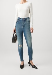 Jeans de tiro alto azules con un lavado medio, con detalles de desgastado en las rodillas, combinados con una blusa blanca de manga larga y tacones negros de punta.