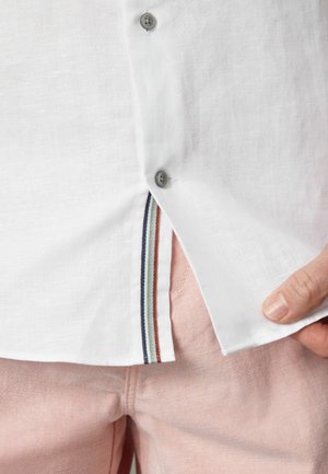 Camisa blanca de botones abierta para revelar detalle de costura interior a rayas sobre pantalones rosa claro.