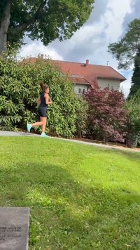 Czarny dopasowany strój sportowy z niebieskimi butami do biegania. Biegacz na ścieżce otoczonej zieloną trawą i krzewami, z domami w tle.