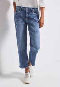 Jeans en denim bleu clair avec une coupe décontractée, taille mi-haute et des patchs déchirés. L'association avec des baskets blanches renforce le look décontracté.