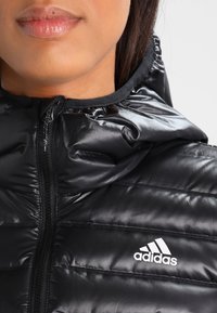 Chaqueta de plumas negra con acabado brillante, que presenta un cuello alto, cremallera frontal y el logo blanco de Adidas en el lado izquierdo.